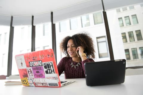A woman on a phone call works across a laptop and tablet in a bright office
